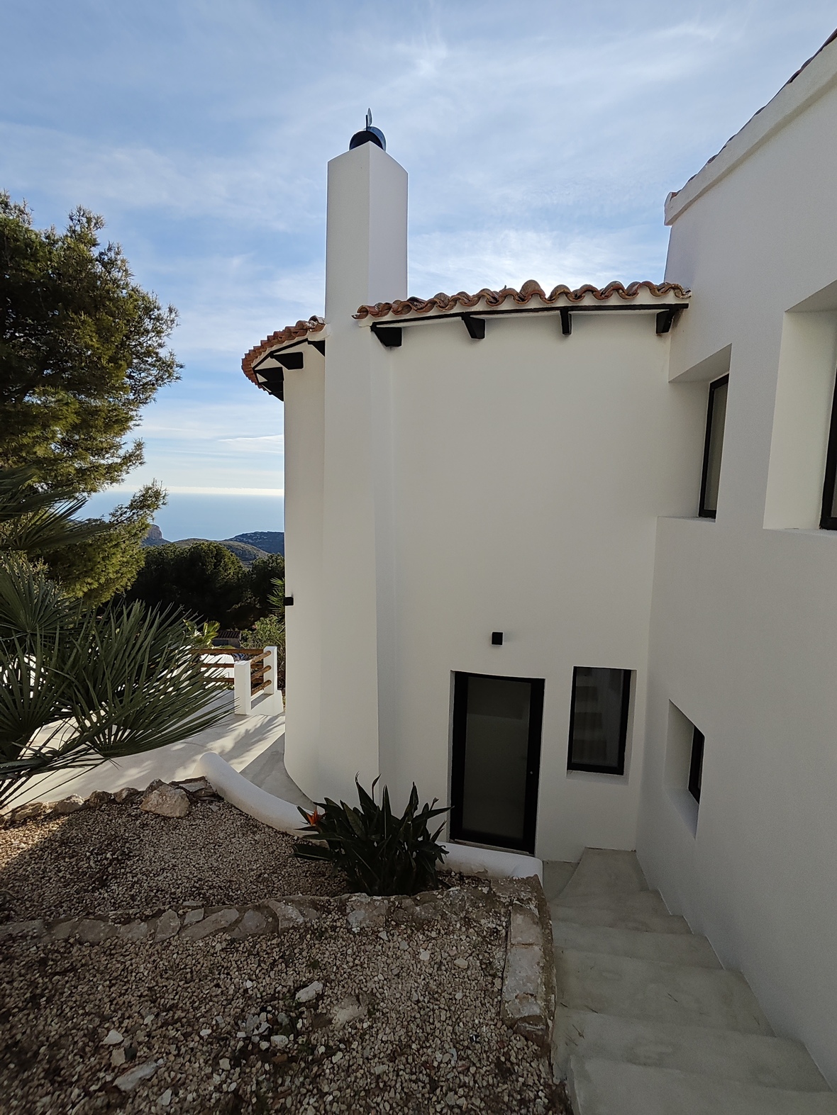 Exterior de villa blanca con chimenea integrada, tejado de terracota y escaleras de piedra que descienden hacia la entrada con vistas al mar.