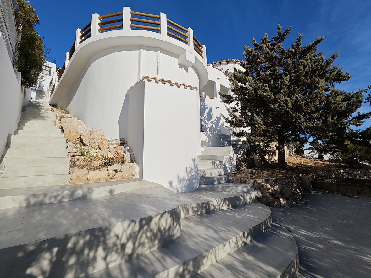Exterior de villa mediterránea con muros curvos blancos, amplias escaleras de hormigón y terraza circular con barandilla de madera natural.