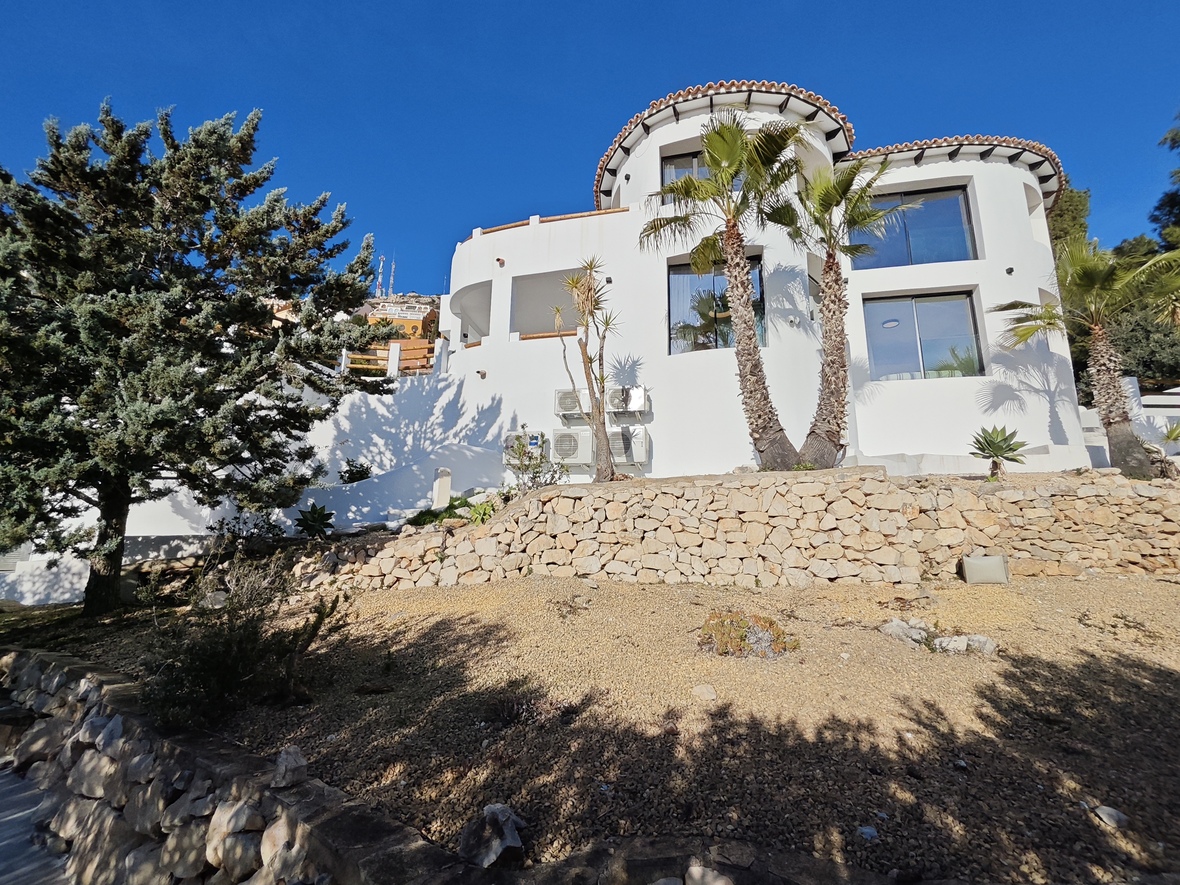 Fachada de villa blanca con arquitectura curva, muros de contención de piedra natural y vegetación mediterránea bajo un cielo despejado.