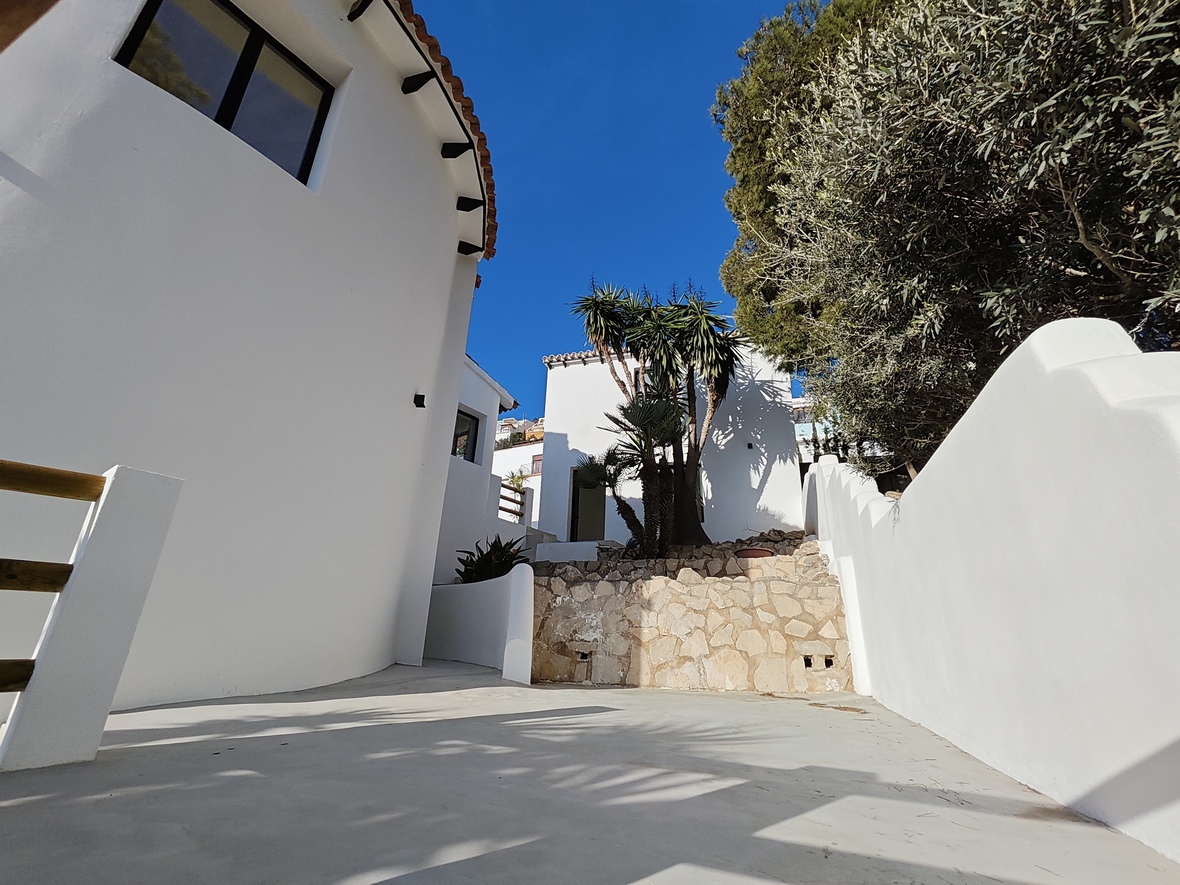 Fachada exterior con paredes blancas curvas, muro de mampostería en piedra natural y zona pavimentada. Incluye vegetación mediterránea con palmeras y árboles maduros.