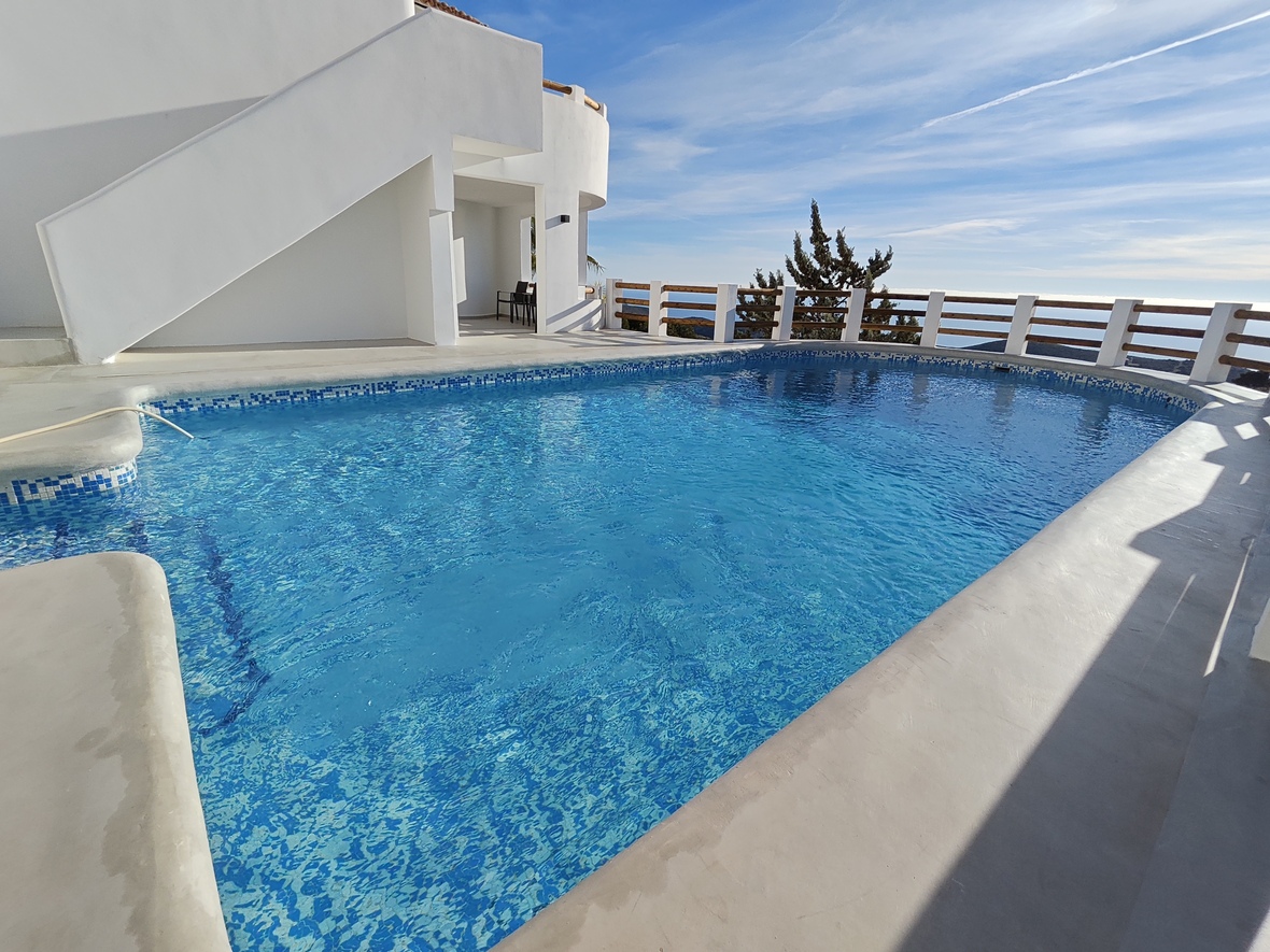 Piscina exterior con revestimiento de gresite azul, terraza de cemento blanco y barandilla de madera con vistas panorámicas al mar.