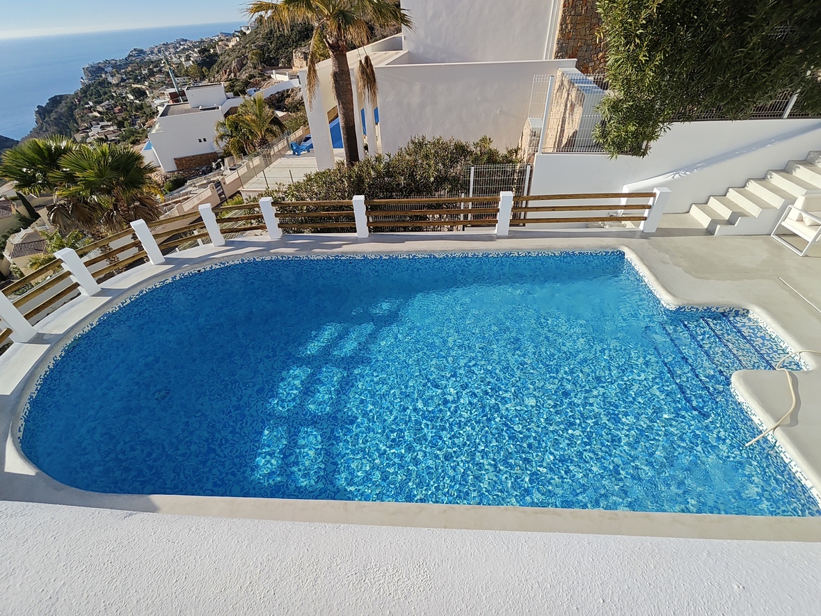 Piscina privada de gresite azul con escaleras integradas, rodeada por una terraza blanca y barandilla de madera con vistas panorámicas al mar Mediterráneo.