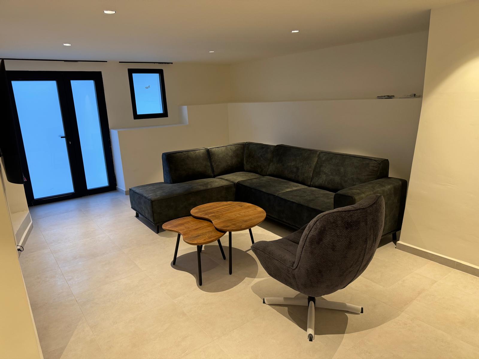 Sala de estar moderna con sofá rinconero gris oscuro, mesas de centro de madera orgánica y sillón giratorio sobre suelo de gres claro con iluminación empotrada.