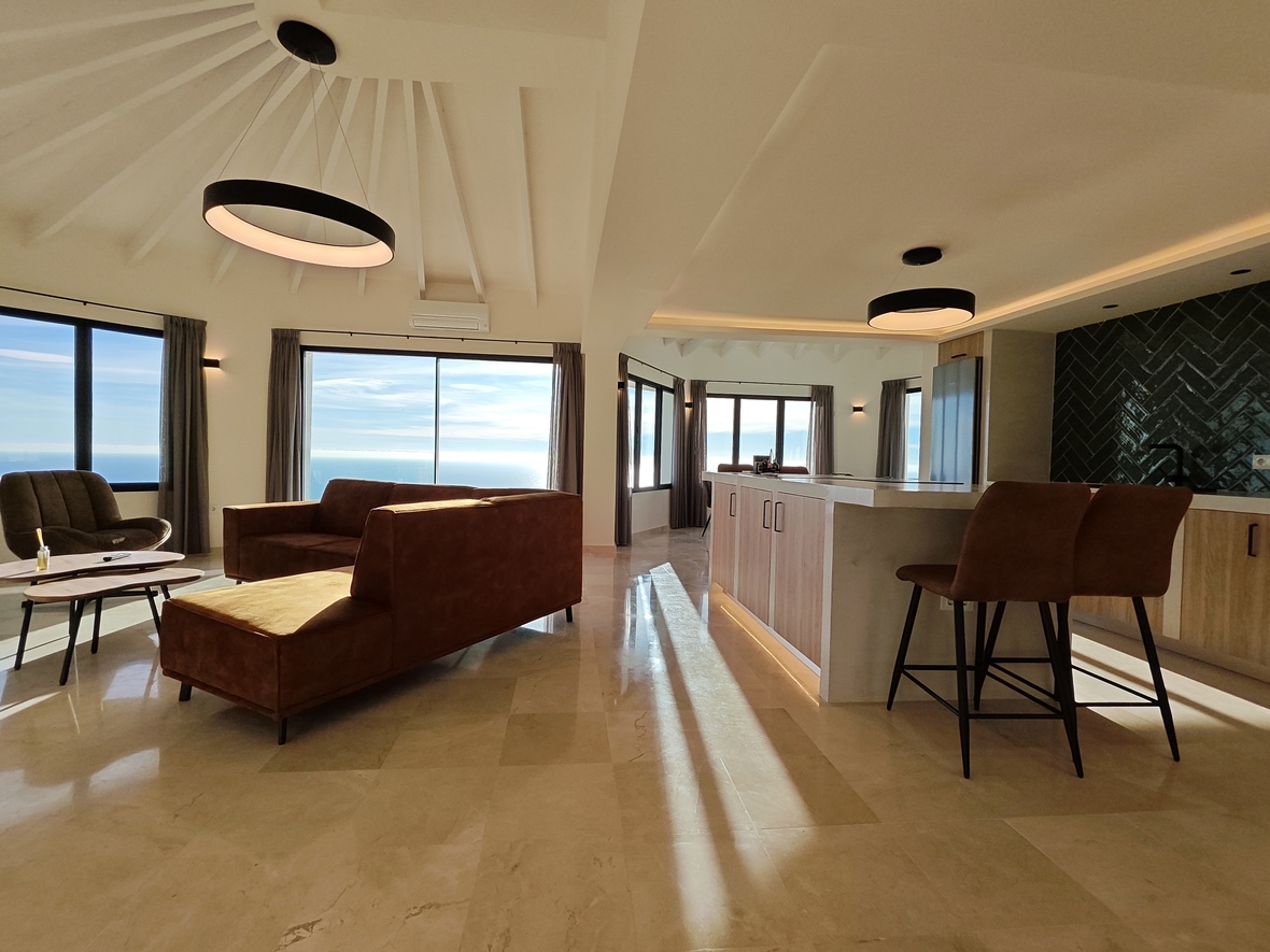 Salón de concepto abierto con techo abovedado de vigas blancas, cocina con isla de mármol y amplios ventanales con vistas panorámicas al océano.