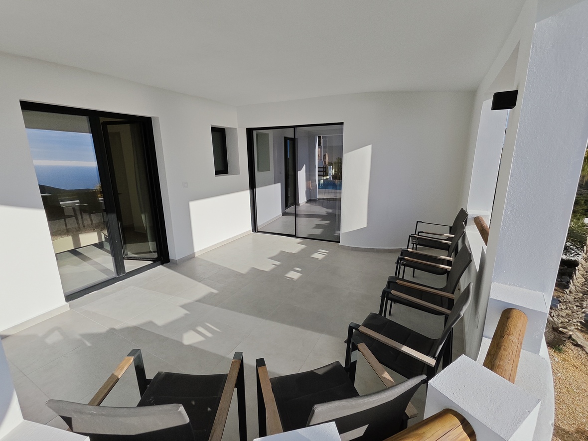 Terraza cubierta con paredes blancas, suelo porcelánico gris claro y cerramientos negros. Incluye sillones modernos con reposabrazos de madera y vistas al mar.