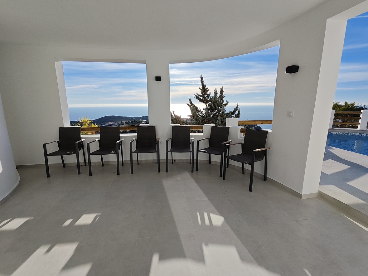 Terraza cubierta con sillones modernos negros, suelo de baldosas grises y amplios ventanales abiertos con vistas panorámicas al mar y acceso a la zona de piscina.