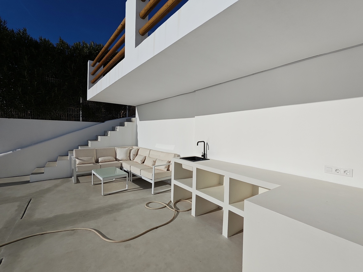 Terraza exterior con cocina de obra blanca, fregadero negro y zona de estar con sofá en L y mesa de centro minimalista bajo la escalera.