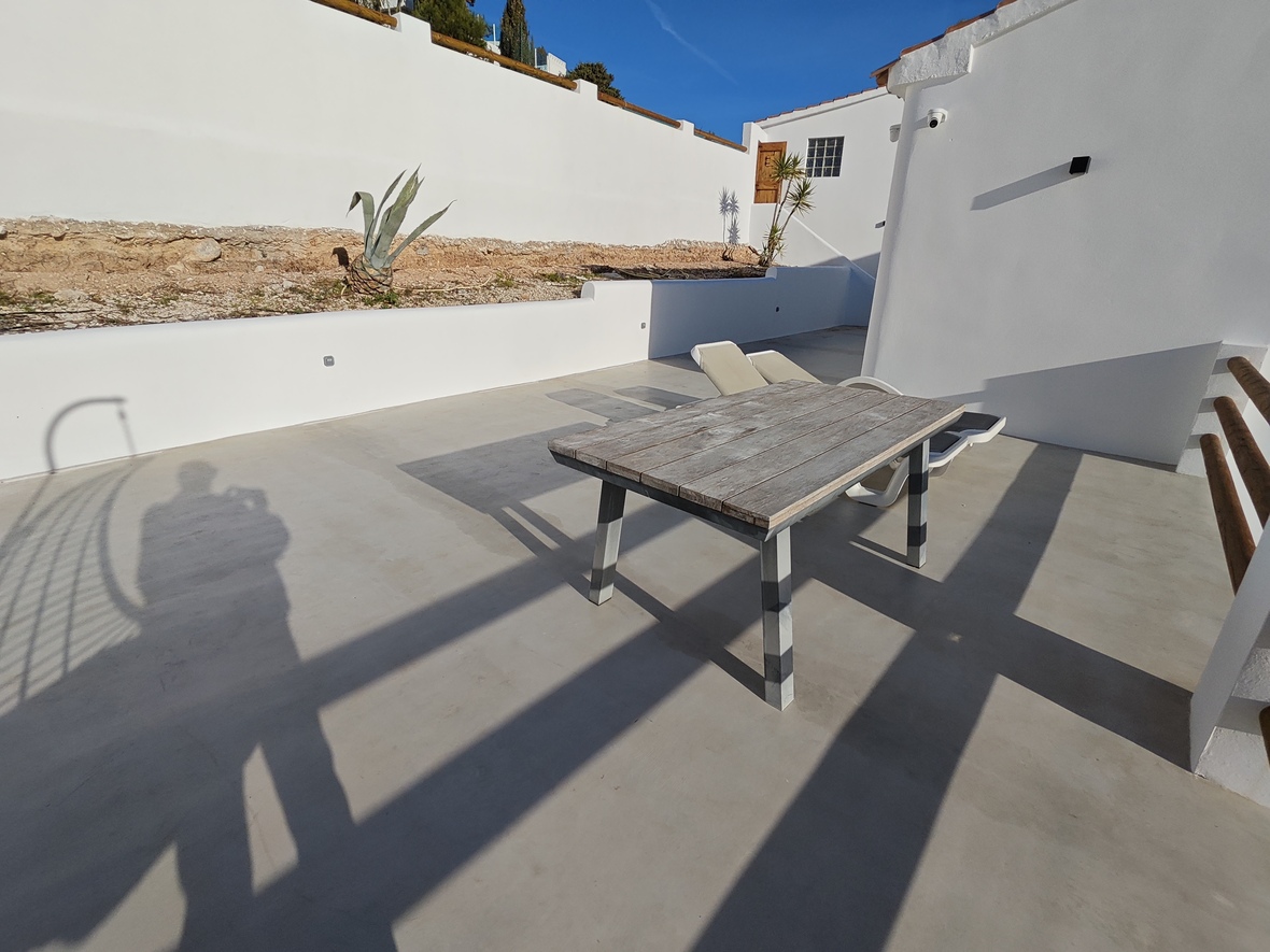 Terraza exterior con suelo de microcemento, mesa de madera rústica y muros blancos bajo un cielo despejado.