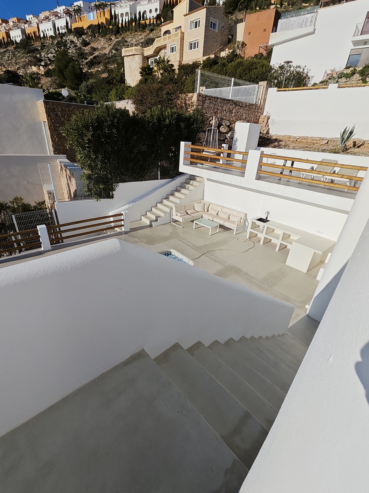 Terraza exterior de varios niveles con paredes blancas, barandillas de madera, zona de estar con sofá en L y cocina exterior integrada frente a una ladera mediterránea.