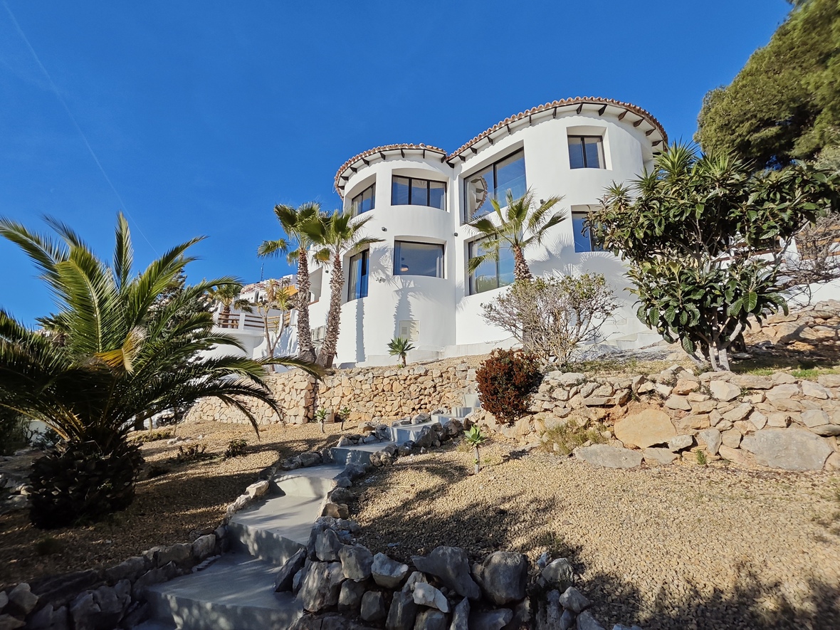 Vista exterior de una villa blanca con líneas arquitectónicas curvas y grandes ventanales. El jardín en pendiente incluye muros de piedra, palmeras y una escalera de hormigón.