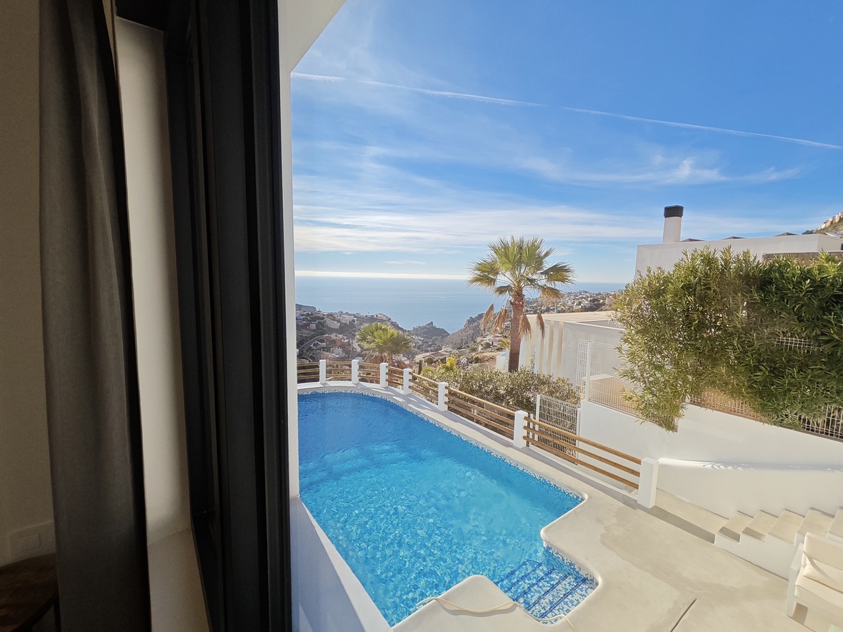 Vista interior hacia una piscina de mosaico azul y terraza blanca con vistas panorámicas a la costa mediterránea y palmeras bajo un cielo despejado.