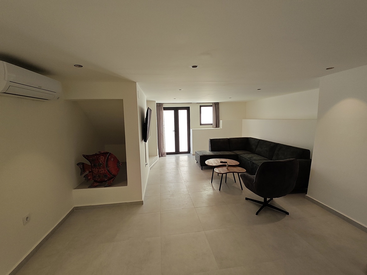 Zona de estar con sofá rinconero gris, mesas de madera y un nicho decorativo con escultura roja. Dispone de aire acondicionado, suelo cerámico y luces empotradas.
