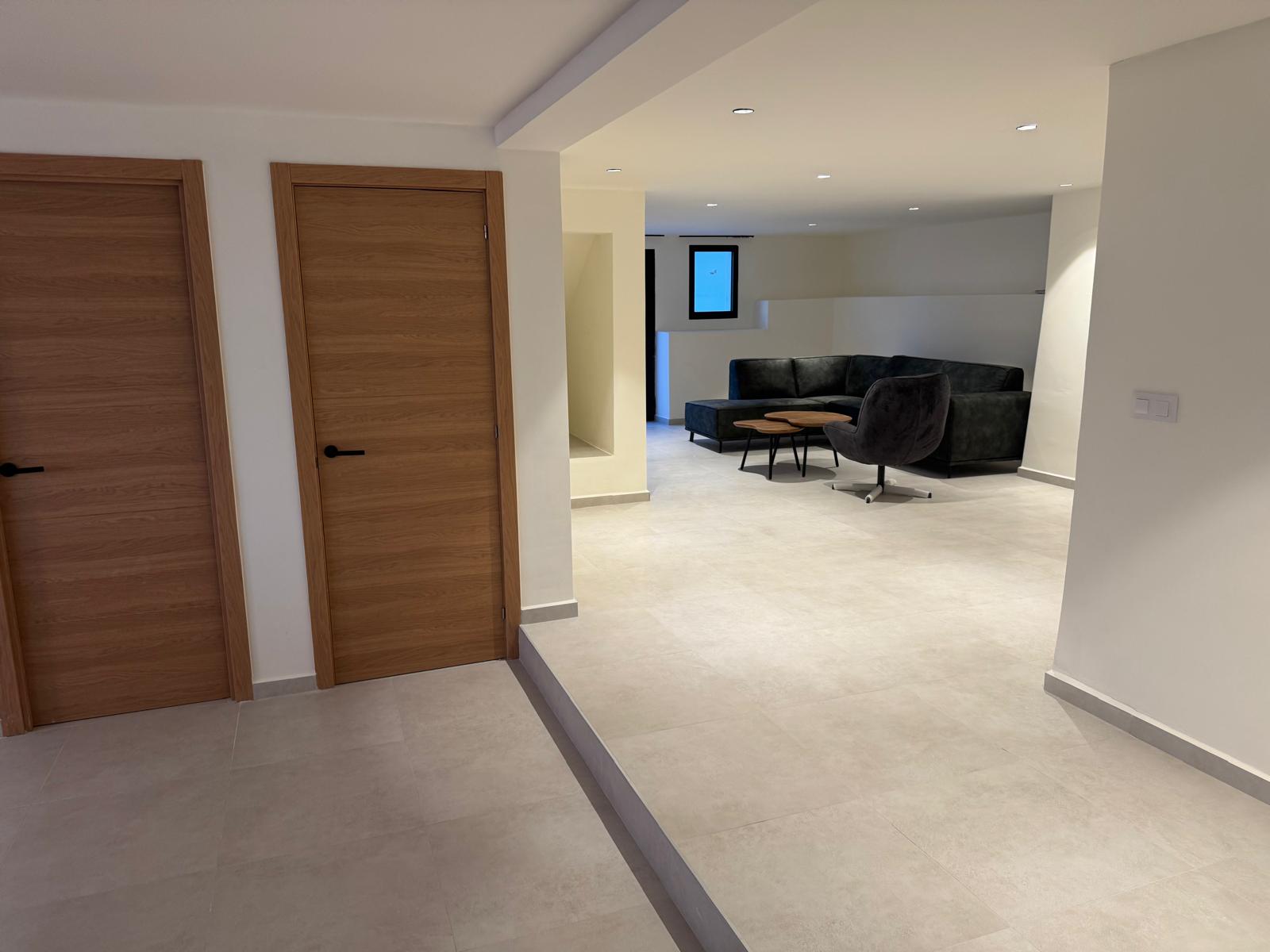 Zona de estar moderna con suelos de gres gris claro, iluminación empotrada y sofá rinconera negro, junto a puertas de madera con veta horizontal.