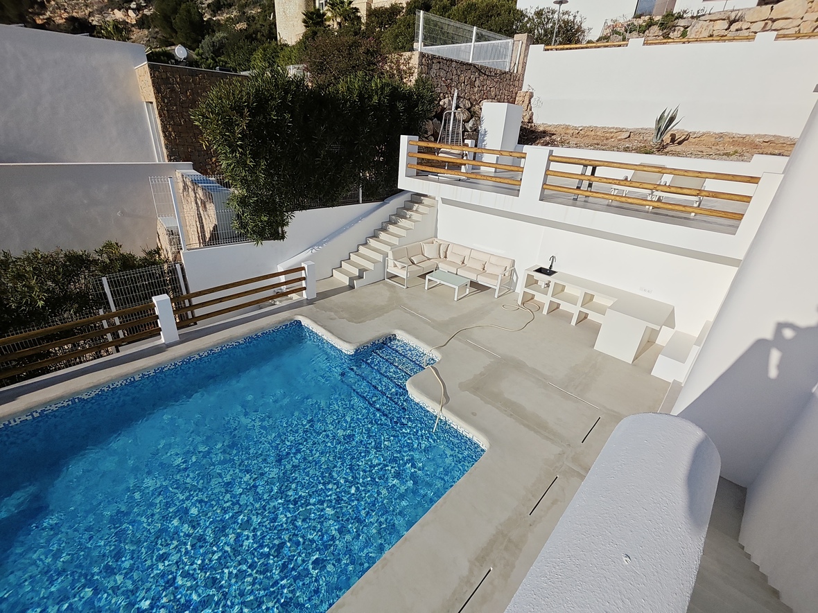 Zona de piscina con solárium de cemento pulido, cocina exterior integrada y terraza superior con barandillas de madera y tumbonas.