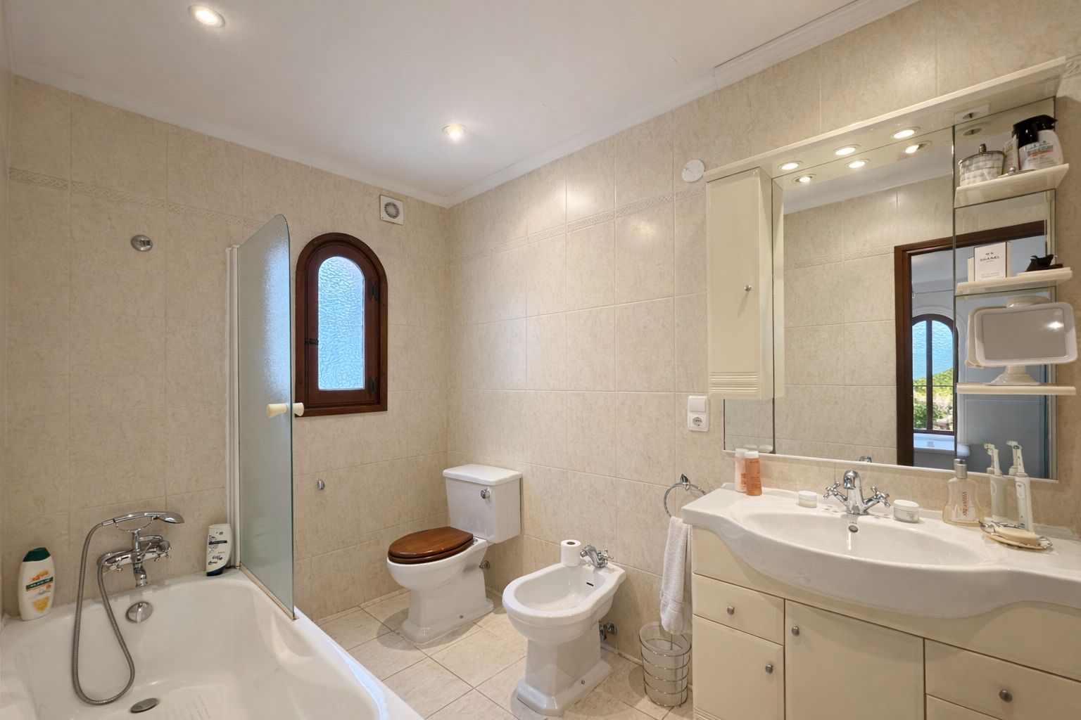 Cuarto de baño con bañera y mampara de cristal, bidé e inodoro con tapa de madera. Incluye mueble de lavabo, espejo con focos y una ventana de arco en madera.
