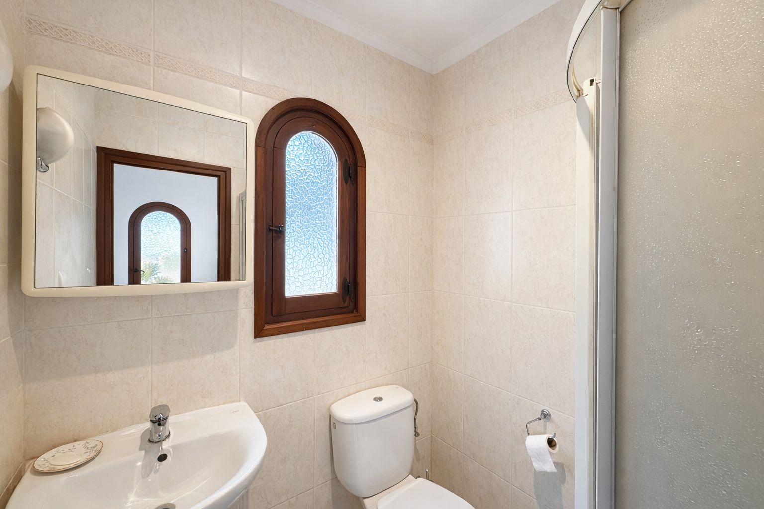 Cuarto de baño con paredes alicatadas en tonos crema, ventana de madera en arco con cristal translúcido y lavabo de cerámica blanca.