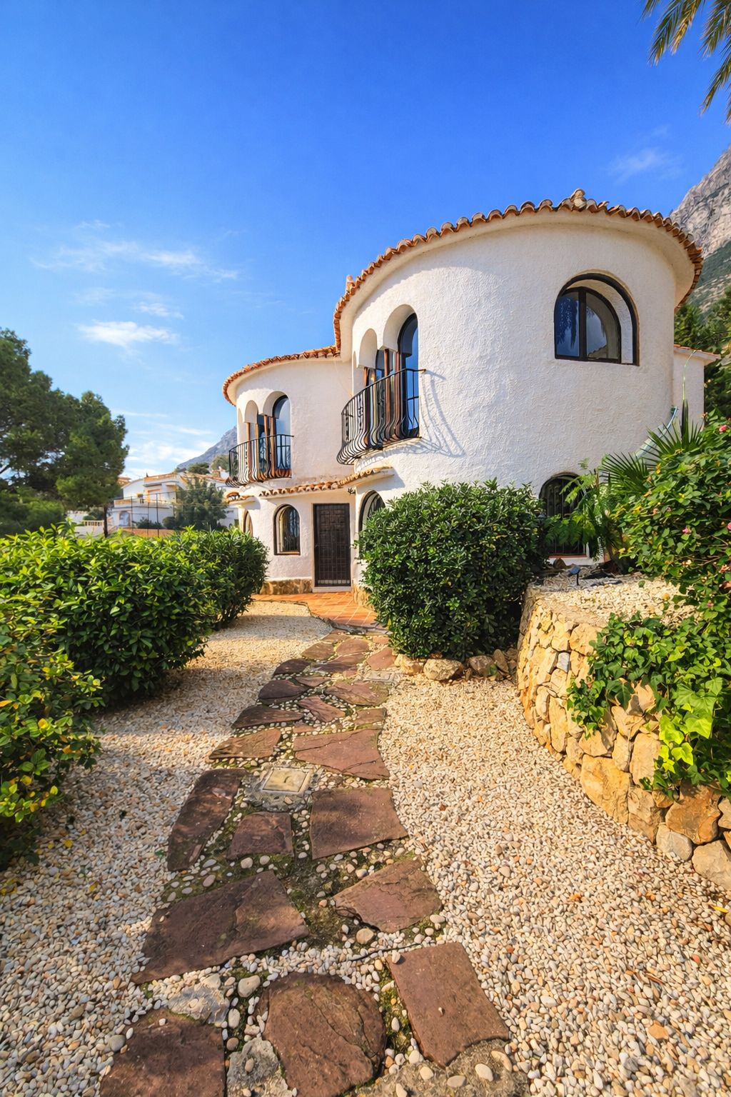Fachada mediterránea con muros curvos blancos, ventanas de arco y balcones de forja. Un camino de piedra natural atraviesa el jardín de grava y arbustos.