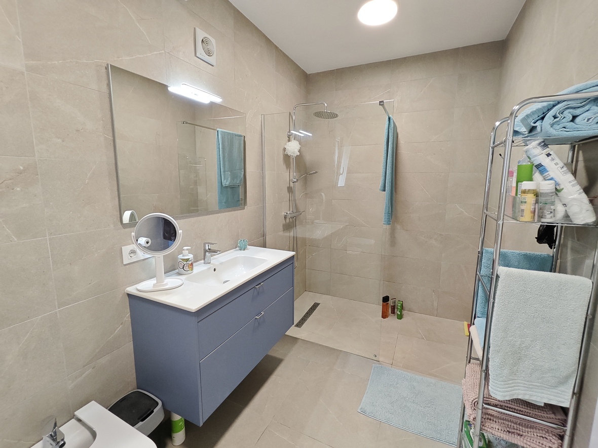 Baño moderno con mueble suspendido azul, amplia ducha a ras de suelo con mampara de cristal y revestimiento cerámico en tonos arena.
