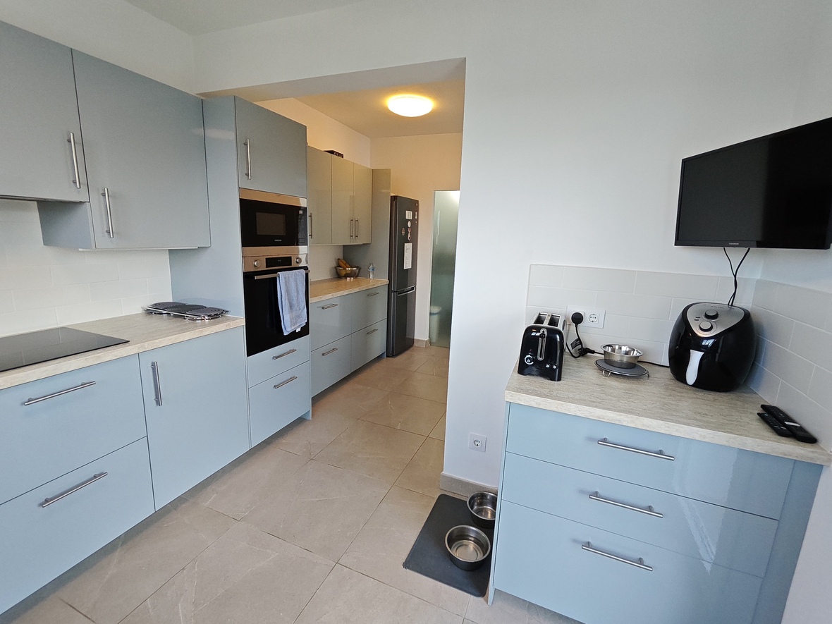 Cocina moderna con armarios azul claro, electrodomésticos integrados y encimeras de efecto piedra. Incluye televisor de pared y suelos de gres gris.