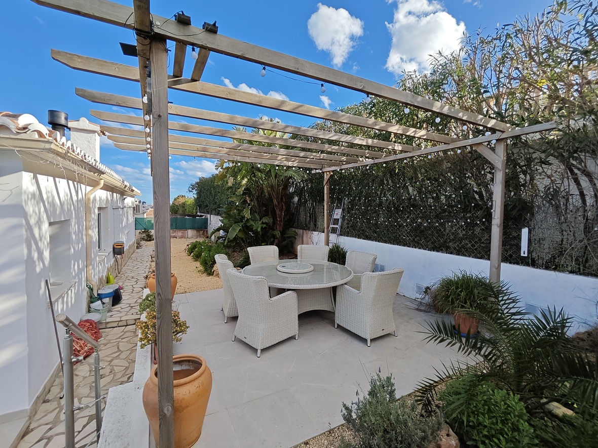 Comedor exterior con mesa redonda de cristal y seis sillas de mimbre blanco bajo una pérgola de madera con guirnaldas de luces y vegetación mediterránea.