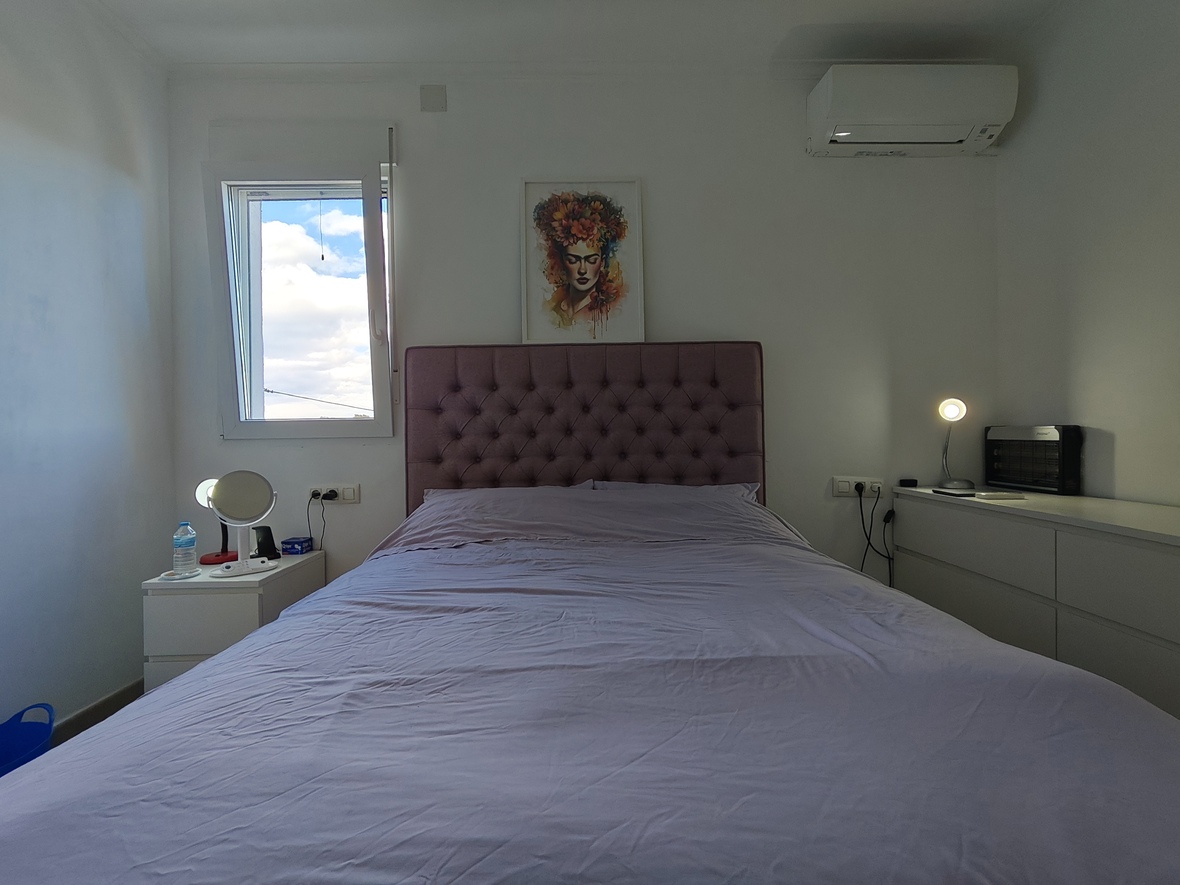Dormitorio con cabecero capitoné en color malva, mobiliario blanco minimalista y aire acondicionado. Incluye un cuadro decorativo y ventana con luz natural.