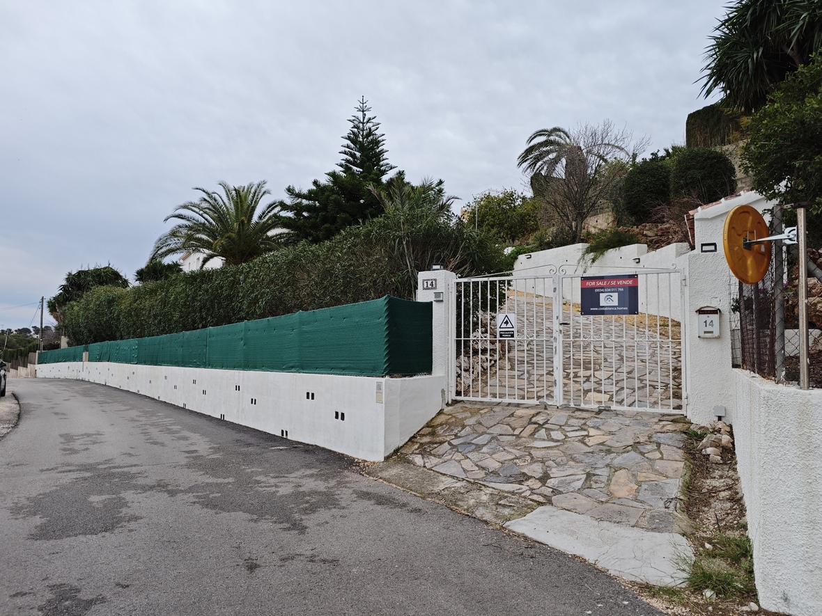 Entrada principal con puerta metálica blanca, rampa de piedra natural y muro perimetral con seto alto y vegetación mediterránea.
