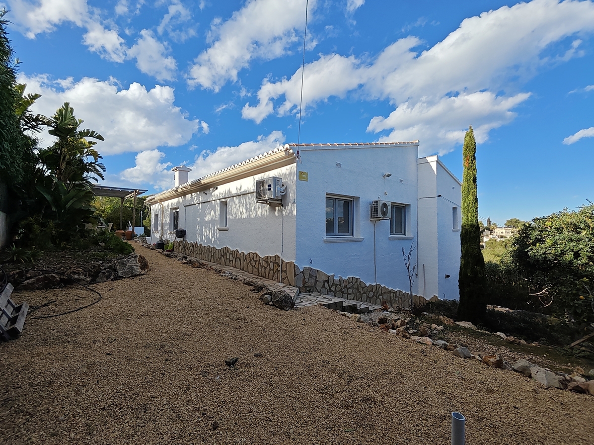 Exterior de la villa con fachada blanca y zócalo de piedra natural, rodeada de vegetación mediterránea y un camino de grava bajo un cielo despejado.