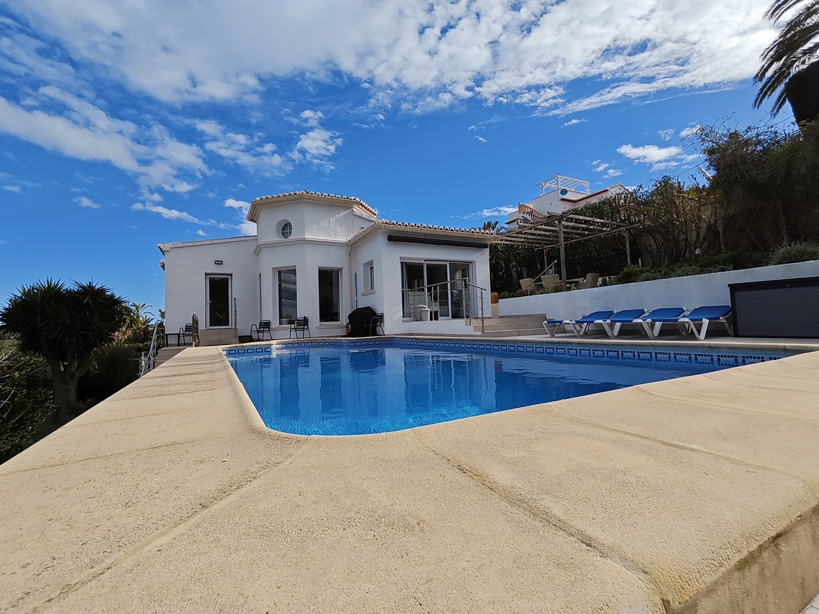 Exterior de villa blanca con piscina privada de gresite azul, terraza de piedra compacta y zona de hamacas junto a la fachada mediterránea.