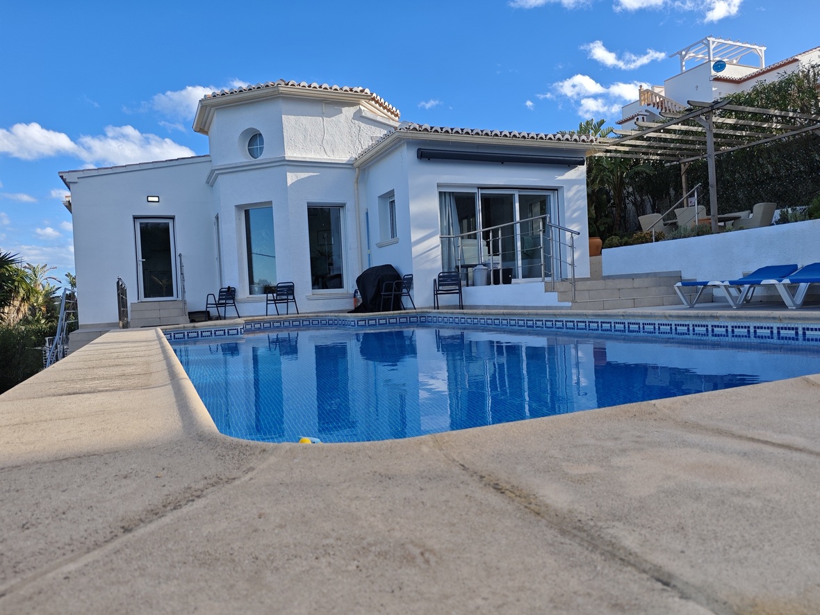 Piscina privada con revestimiento de gresite azul y terraza de piedra, situada frente a una villa blanca de estilo mediterráneo con torreón octogonal y ventanal circular.
