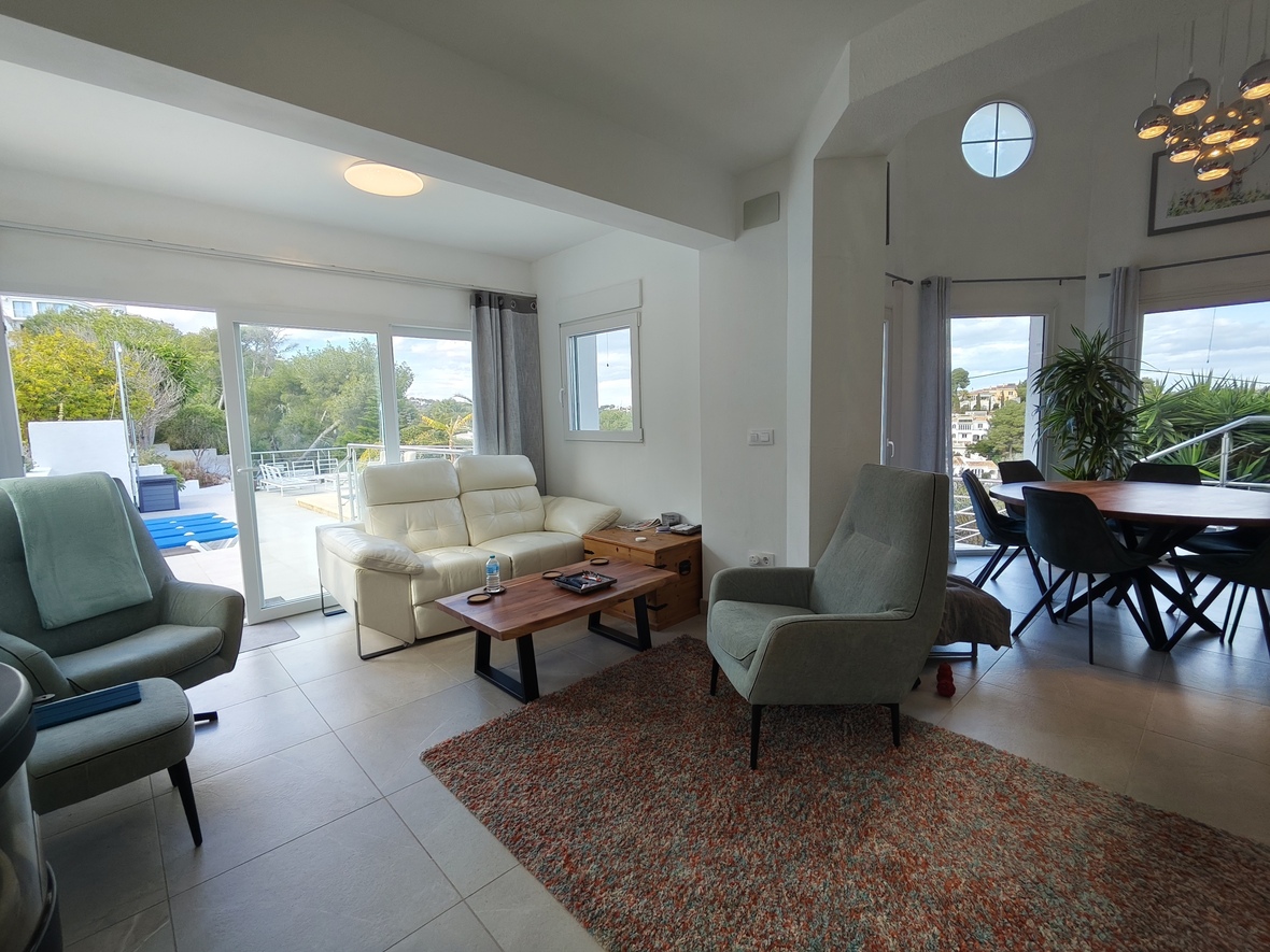 Salón comedor de planta abierta con sofá de cuero blanco, techos altos y ventanales de suelo a techo que conectan con la terraza y la zona de piscina.
