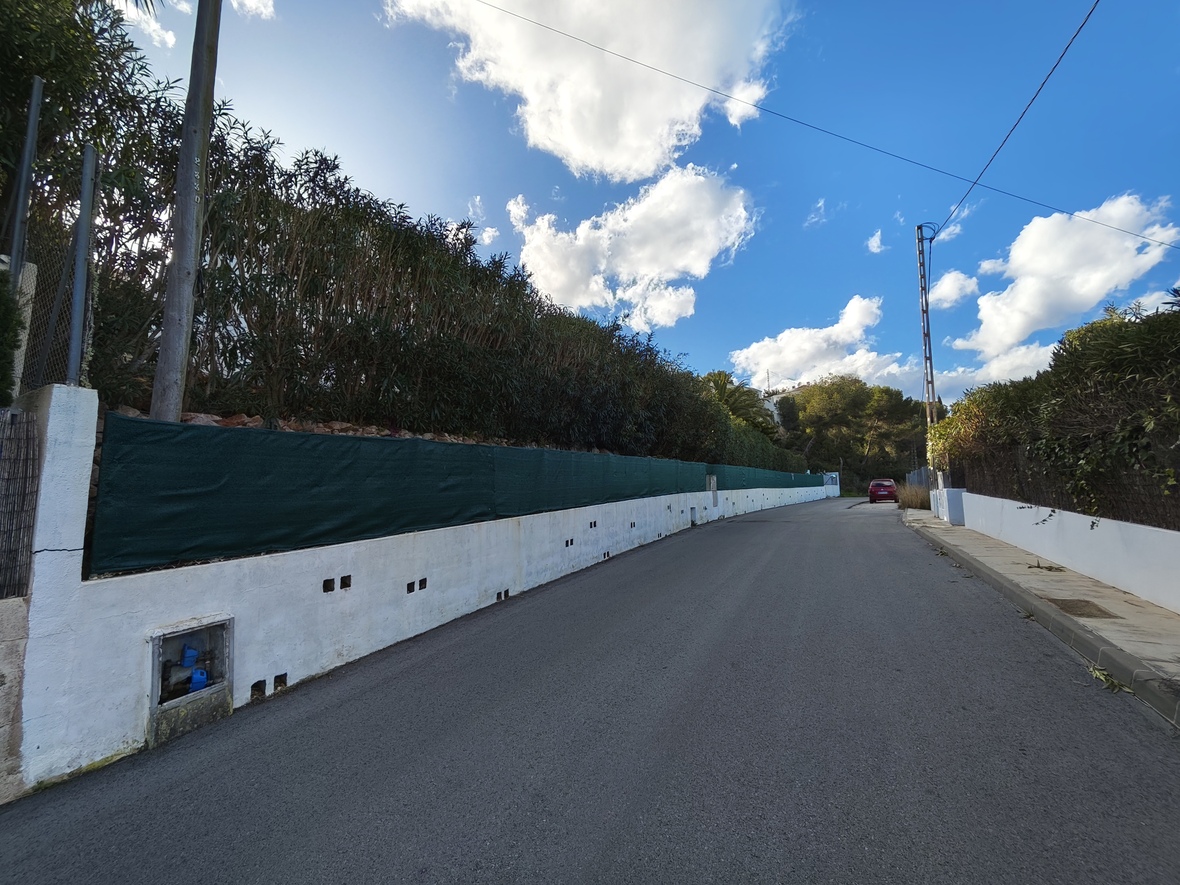 Vista de la calle de acceso con muro perimetral blanco, malla de privacidad verde y vegetación densa bajo un cielo azul con nubes blancas.