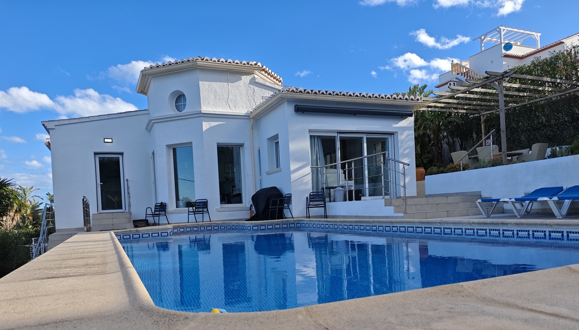Vista exterior de una villa mediterránea blanca con piscina privada, terraza con tumbonas azules y una pérgola de madera bajo el cielo despejado.
