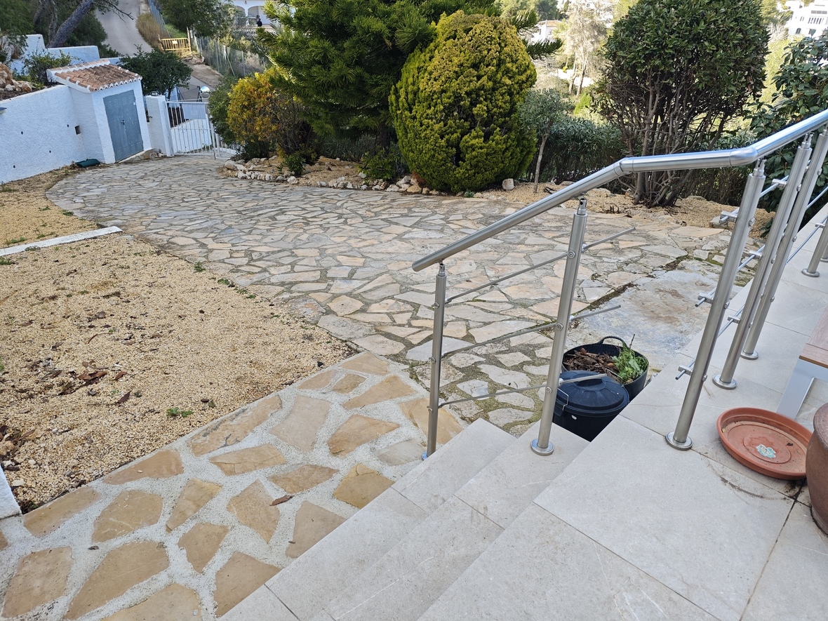 Vista exterior que muestra un camino de piedra natural rodeado de vegetación mediterránea y una escalera con barandilla de acero inoxidable.