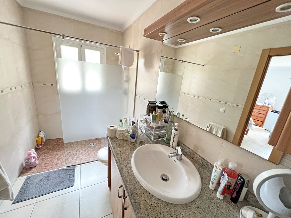 Baño con encimera de granito, lavabo encastrado y mueble de madera. Dispone de ducha con mampara de cristal translúcido y suelos cerámicos en tonos beige.