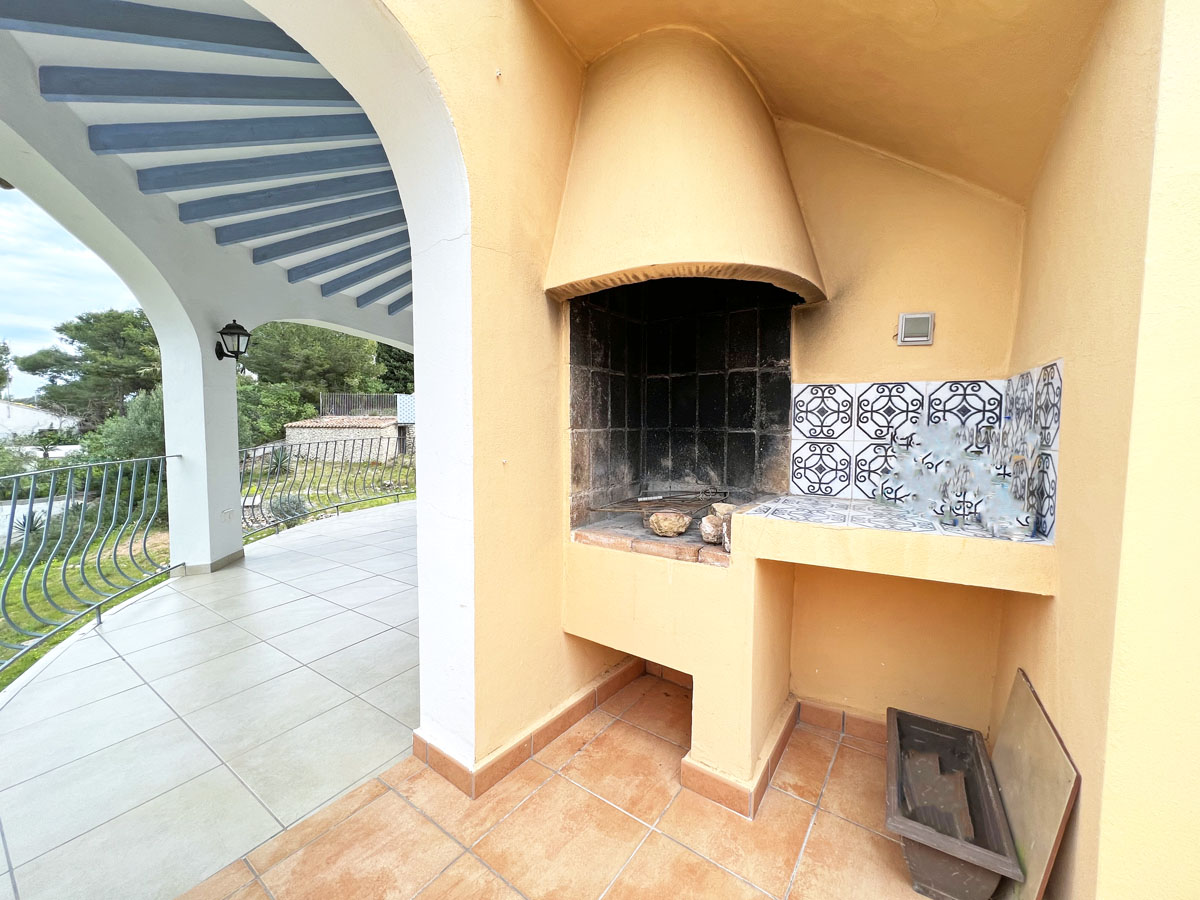 Barbacoa de obra con campana cónica y encimera de azulejos tradicionales, situada en una terraza cubierta con arcos blancos y vigas vistas en azul.