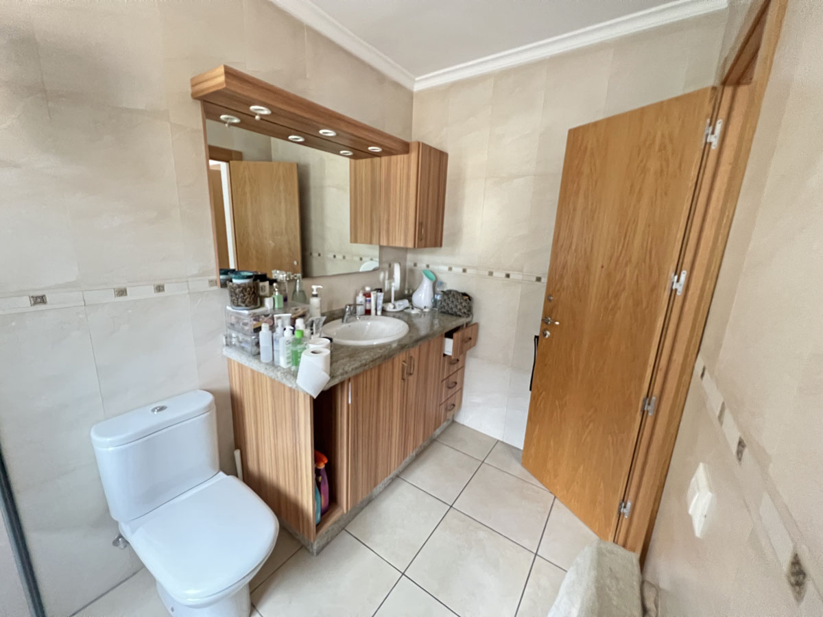 Cuarto de baño con mueble de lavabo en madera, encimera de granito y espejo con iluminación integrada. Paredes alicatadas en tonos crema con cenefa decorativa.