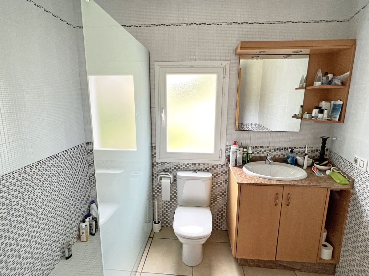 Cuarto de baño funcional con ducha de obra, mampara de cristal translúcido, mueble de lavabo en madera y paredes revestidas con azulejos de mosaico decorativo.
