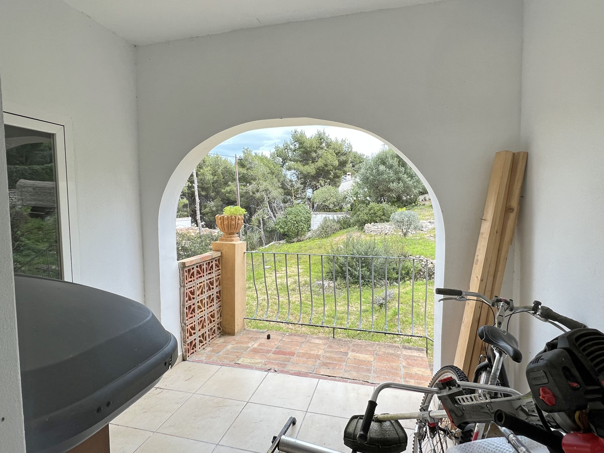 Porche cubierto con arco de medio punto que ofrece vistas al jardín mediterráneo y pinada, con suelos cerámicos y barandilla metálica.