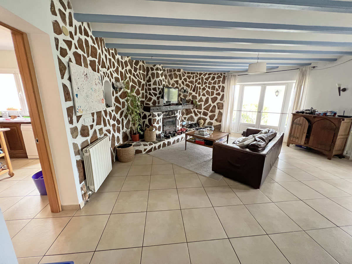 Salón rústico con pared de piedra natural, chimenea integrada y vigas de madera en azul claro sobre suelos cerámicos con acceso al exterior.