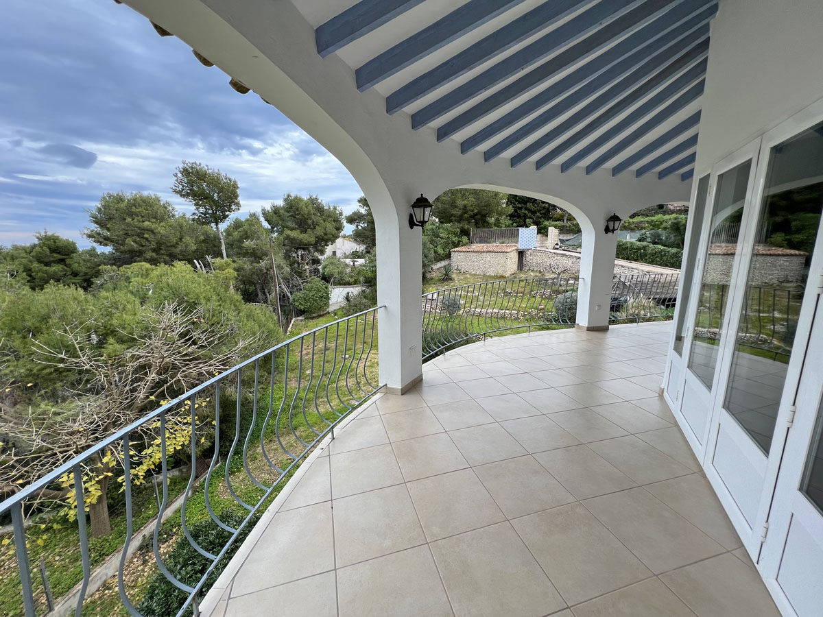 Terraza cubierta con arcos, vigas de madera en azul pálido y suelo cerámico. Ofrece vistas al jardín mediterráneo y cuenta con una barandilla metálica curva.