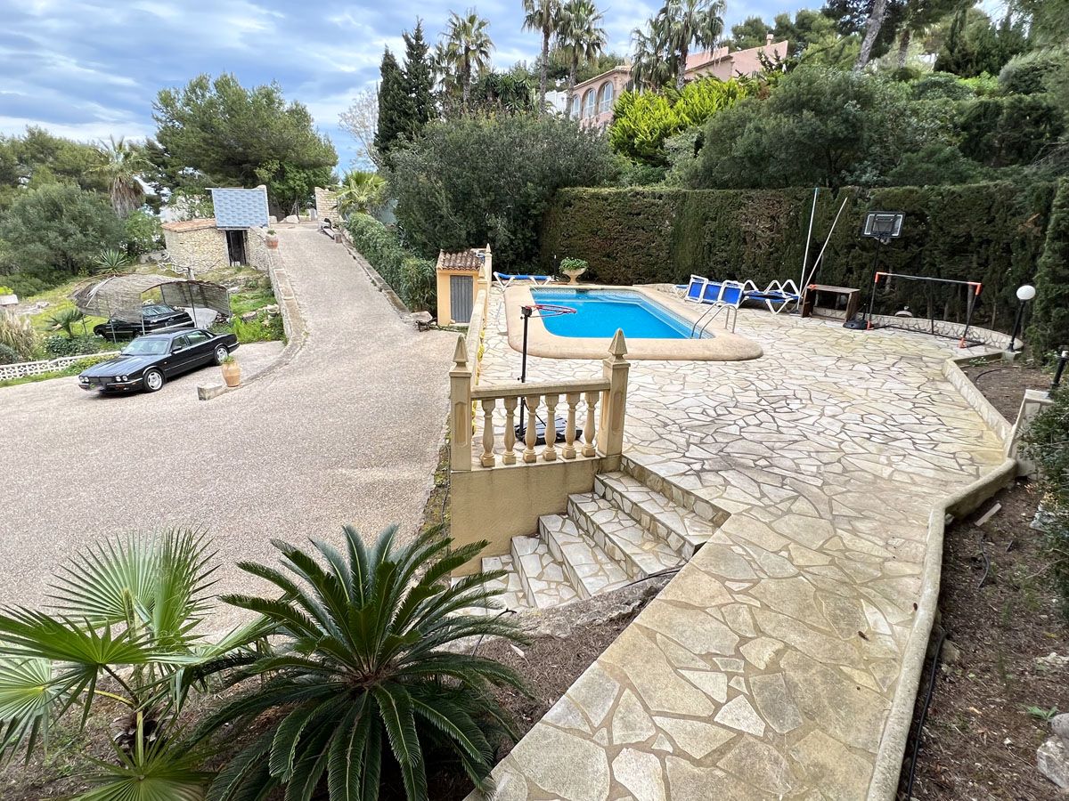 Terraza elevada con piscina de 8x4 metros, pavimentada en piedra natural y rodeada de vegetación mediterránea, con acceso directo a la zona de aparcamiento inferior.