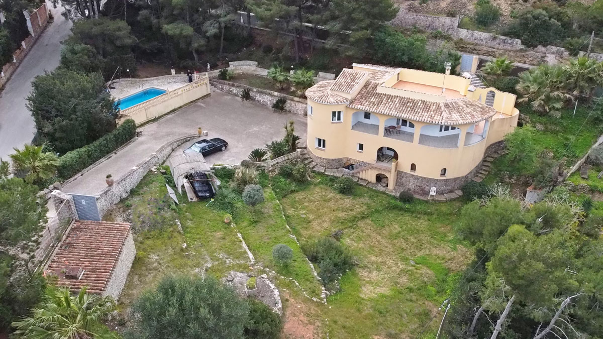 Vista aérea de la villa con fachada mediterránea, naya arqueada y base de piedra natural. Incluye amplia zona de aparcamiento, piscina privada y jardín en bancales.