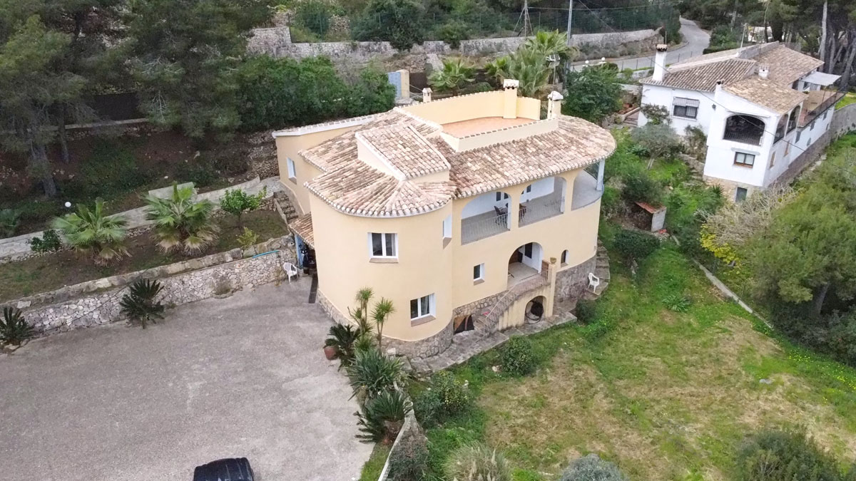 Vista aérea de la villa mediterránea con fachada curva, tejado de teja cerámica y amplia zona de aparcamiento pavimentada rodeada de vegetación autóctona.