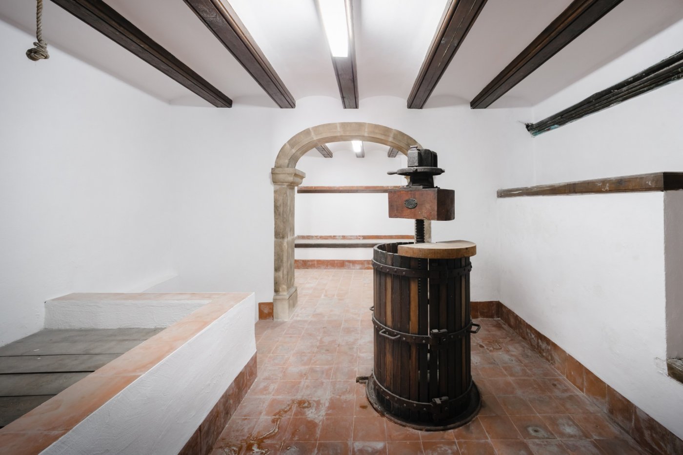 Bodega tradicional con prensa de vino de madera, suelos de barro cocido, vigas de madera oscura y un arco de piedra tosca original.