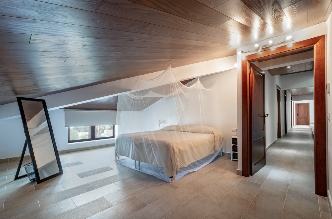 Dormitorio abuhardillado con techos de madera, cama con mosquitera y suelos cerámicos. Incluye un espejo de pie y acceso a un pasillo con carpintería de madera natural.