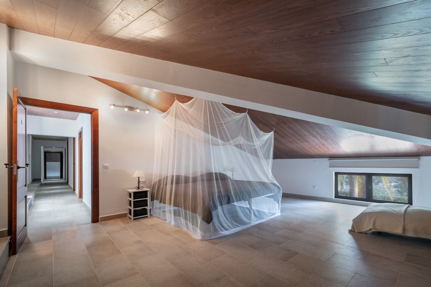 Dormitorio abuhardillado con techos de madera natural, suelos cerámicos y cama con mosquitera. La estancia conecta con un pasillo distribuidor y ofrece vistas al jardín.