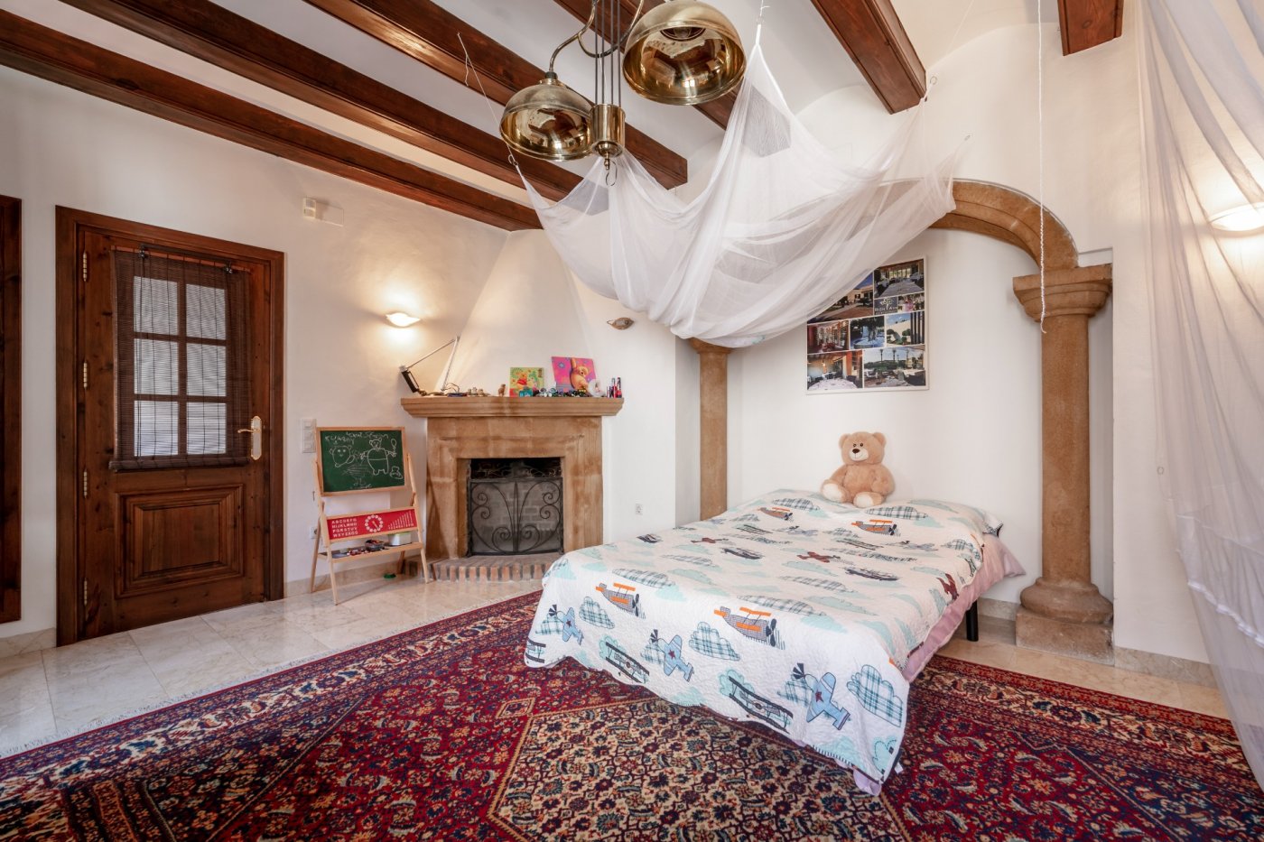 Dormitorio con vigas de madera, chimenea de piedra tosca y arco arquitectónico, complementado con suelos de mármol y una alfombra de diseño tradicional.