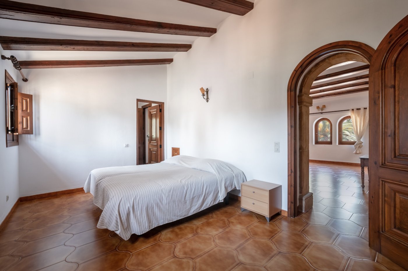 Dormitorio de estilo mediterráneo con vigas de madera a la vista, suelos de gres rústico y un arco de piedra tosca que comunica con las estancias contiguas.