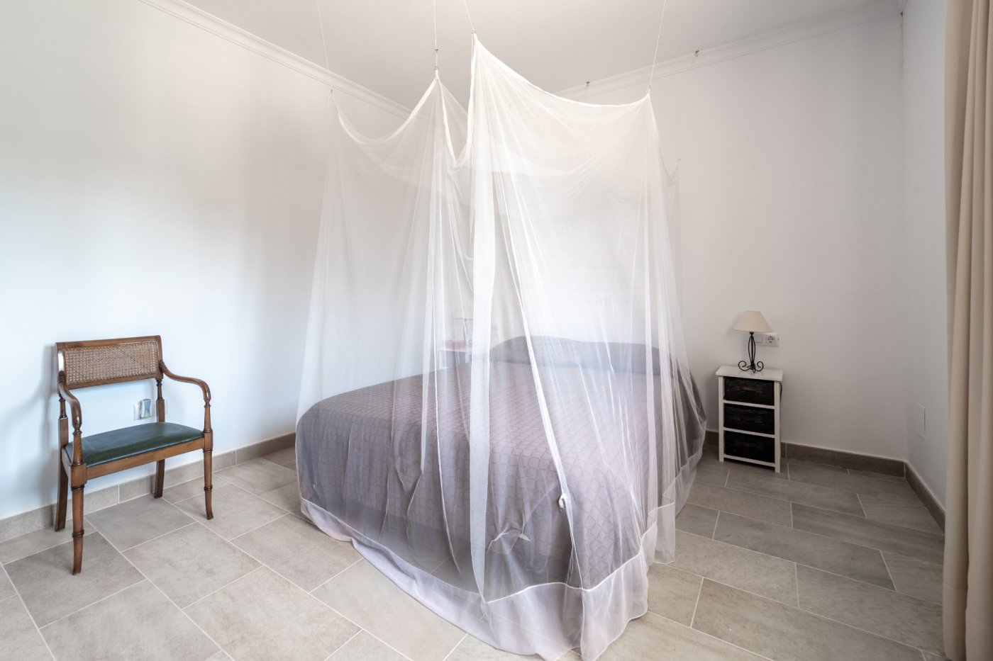 Dormitorio luminoso con suelos de gres porcelánico, cama doble con mosquitera de techo y mobiliario de estilo clásico sobre paredes blancas.