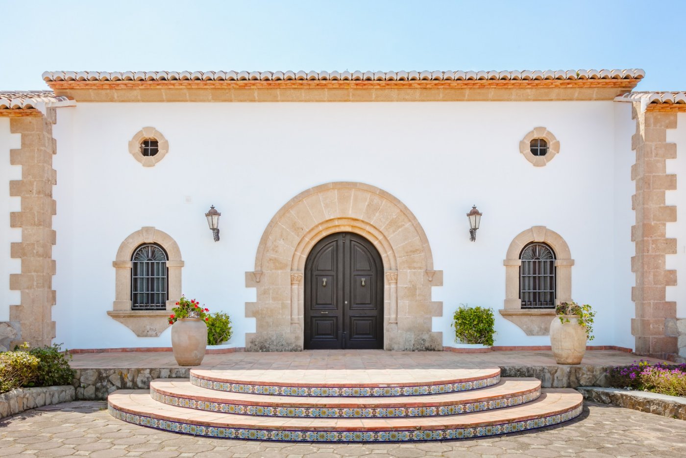 Entrada principal con arco de piedra tosca, puerta de madera oscura y escalinata semicircular con azulejos decorativos sobre fachada blanca tradicional.